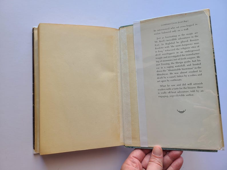 May include: An open book with aged, yellowed pages. The right page has printed text in black, with a small illustration at the bottom. The left page is blank. The book is held open by a hand.