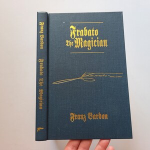 Puede incluir: Un libro de tapa dura de color azul oscuro con letras doradas titulado "Frabaato el Mago" de Franz Bardon. El libro tiene una firma dorada en la portada.