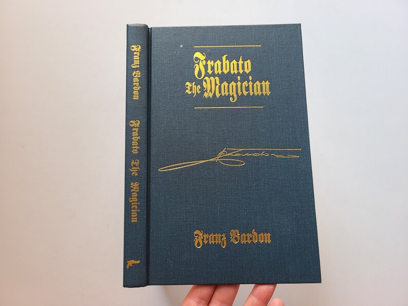 Puede incluir: Un libro de tapa dura de color azul oscuro con letras doradas titulado "Frabaato el Mago" de Franz Bardon. El libro tiene una firma dorada en la portada.