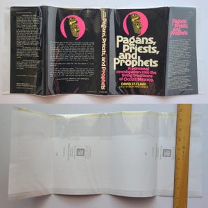 Pu&ograve; includere: Copertina del libro "Pagans, Priests, and Prophets" di David St. Clair. La copertina &egrave; nera con testo rosa e bianco. Presenta una grafica di un volto all'interno di un cerchio rosa. Il libro &egrave; in una custodia di plastica trasparente.