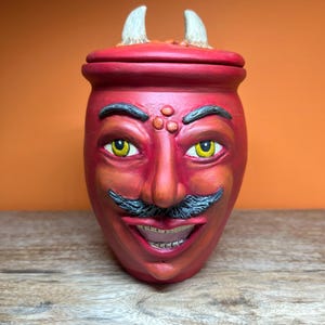 May include: A red ceramic jar with a lid featuring two white horns. The jar is sculpted with a face that has yellow eyes, a black mustache, and a wide, smiling mouth showing teeth. The surface has a matte finish with subtle shading.
