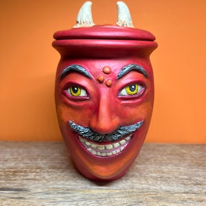 May include: A red ceramic jar with a lid featuring two white horns. The jar is sculpted into a face with yellow eyes, a grey mustache, and a wide grin showing teeth. There are three small orange dots on the forehead between the dark eyebrows.