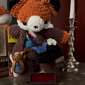 May include: A crocheted fox plush toy wearing a brown coat, blue vest, and red tie. It carries a black satchel with a blue butterfly accessory and playing cards, a brown leather potion bottle holder, and stands behind a small black velvet-lined box.