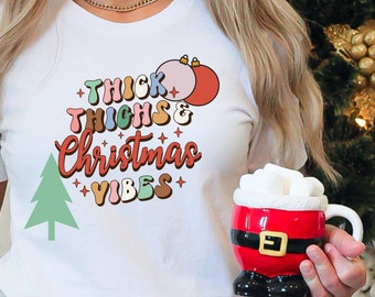 Puede incluir: Camiseta blanca con un gráfico colorido que dice "Thick Thighs & Christmas Vibes". El gráfico incluye un árbol de Navidad y adornos.