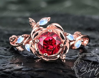 May include: A rose gold ring with a red gemstone set in a rose-shaped band. The ring is adorned with delicate leaves and white gemstones.