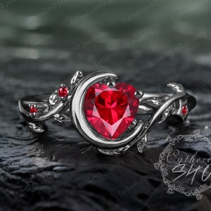 以下が含まれることがあります： ハート型の赤い宝石が特徴のシルバーリング。三日月デザインにセットされ、繊細な葉とツルのディテール、小さな赤い宝石で飾られています。このリングはジュエリーです。