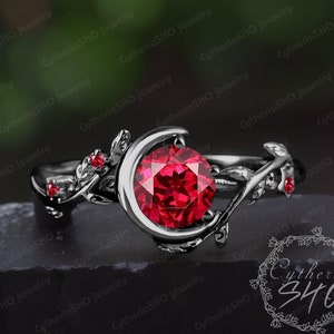 May include: A black ring with a crescent moon design and a large red gemstone. The ring is set with smaller red gemstones on the band and has a vine-like design.