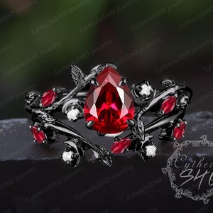 May include: A black ring with a pear-shaped red gemstone surrounded by smaller red gemstones and white diamonds. The ring features a vine design with leaves and branches.