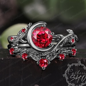 May include: A black metal ring set featuring a ruby gemstone and smaller red accent stones. The engagement ring has a crescent moon design, and the wedding band has a leaf pattern. The rings are set on a dark surface.