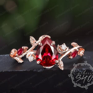 以下が含まれることがあります： バラ色のゴールドにセットされた洋梨型のルビーの宝石のリング。繊細な蔦のデザインで、葉と小さなダイヤモンドが飾られています。リングは黒い背景の上にあります。