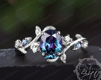 May include: A silver ring featuring a large oval alexandrite gemstone, surrounded by smaller diamonds and alexandrite stones. The ring has a delicate vine design with leaves and branches.