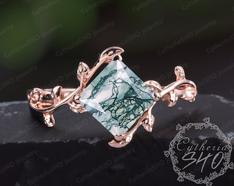 Puede incluir: Un anillo de oro rosa con una piedra preciosa cuadrada de ágata verde musgo engastada en un delicado diseño de vid. El anillo está sobre una superficie negra con el texto "Cytheria 340" visible.