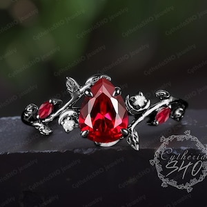 May include: A black ring with a large, pear-cut, bright red gemstone. The ring has a floral design with smaller red stones and white accents. The ring is displayed against a dark backdrop, emphasizing the intricate details of the jewellery.