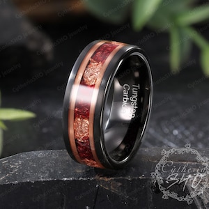 May include: A black tungsten carbide ring featuring a central inlay of crushed red stone and copper-colored flakes. The interior of the band is clearly engraved with the text Tungsten Carbide. The ring has a polished finish and a flat profile.