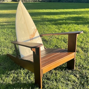 May include: A wooden Adirondack chair with a surfboard leaning against it. The chair is made of dark brown wood and has wide armrests. The surfboard is off-white with orange text that reads "A. Patterson". The chair is on a grassy lawn.