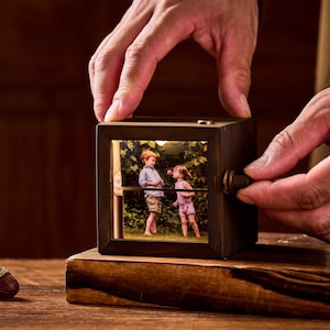 May include: A dark wooden photo cube with a picture of two children. The cube has a small knob on the side. The cube is being held by a person's hands.