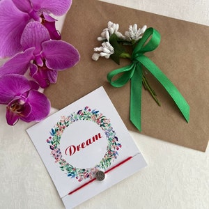 Puede incluir: Una composición que presenta una orquídea púrpura, un sobre de papel kraft y una tarjeta blanca con la palabra "Dream" en escritura roja. También hay una pulsera de hilo rojo con un dije plateado y un pequeño ramo floral con una cinta verde.