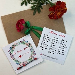 Puede incluir: Una tarjeta de felicitación con la frase "I love you" y una tabla de código Morse. Se muestra una pulsera con cuentas rojas y negras. También hay una flor roja y una cinta verde.