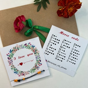 Puede incluir: Una pulsera hecha a mano con cuentas negras y naranjas, presentada con una tarjeta que dice "I love you" y una tabla de código Morse. La tarjeta está decorada con una corona floral. Una flor roja y una cinta verde también están en la imagen.