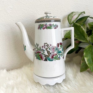 May include: White porcelain teapot with a floral design in shades of pink, green, and gold. The teapot has a round body and a spout on the side. It sits on a white pedestal base.