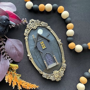 May include: A Halloween-themed decorative piece featuring a haunted house design within an ornate, gold-colored frame. The house is gray with a yellow-lit window and a purple door. A black raven and decorative beads are also present.