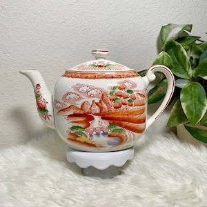 May include: A white teapot with a brown, green, and blue landscape design. The teapot has a round body and a spout on the left side. The teapot is sitting on a white pedestal.