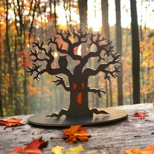 May include: A black wooden Halloween tree cutout with an owl and a jack-o'-lantern face. The tree is sitting on a wooden surface with fall leaves scattered around it.