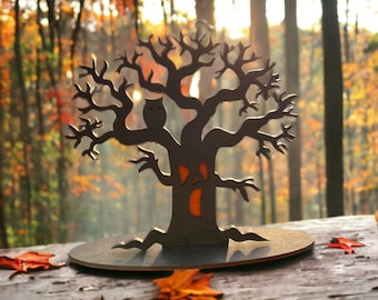 Può includere: Un albero di Halloween in legno nero con un gufo e un viso di zucca. L'albero è appoggiato su una superficie di legno con foglie autunnali sparse intorno.