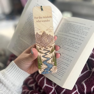 May include: Wooden bookmark with a mountain and river landscape design. The bookmark has the text "For the readers who wander". The bookmark is held open in a book.