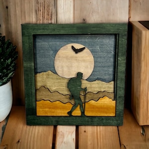 May include: A green wooden frame with a layered wood art piece inside. The art piece depicts a hiker with a backpack walking through a mountainous landscape with a full moon in the sky. The hiker is a silhouette, and the moon is a light brown color.