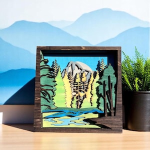 May include: A wooden framed wall art piece depicting a mountain scene with a river and trees. The mountain is light gray, the trees are green and yellow, and the river is blue.