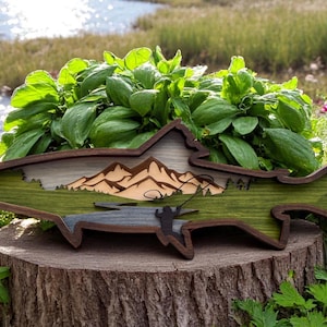 K&ouml;nnte beinhalten: Wandkunst aus Holz in Form einer Forelle mit einer Bergszene und einem Fischer, der eine Leine wirft. Die Forelle ist in Gr&uuml;n- und Braunt&ouml;nen bemalt.