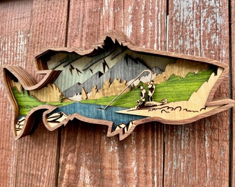 Può includere: Un'opera d'arte in legno a strati a forma di pesce, raffigurante una scena di pesca. L'opera presenta montagne, un fiume e due figure che pescano. La tavolozza dei colori comprende sfumature di marrone, verde, blu e grigio.