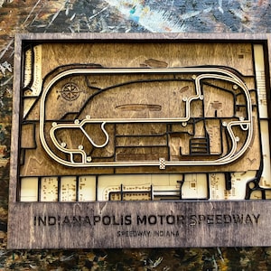 May include: A wooden 3D model of the Indianapolis Motor Speedway race track. The track is made of light brown wood and is surrounded by a dark brown frame. The text "Indianapolis Motor Speedway" and "Speedway, Indiana" is printed below the track.