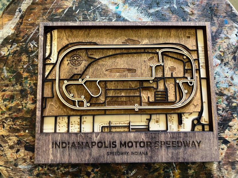 May include: A wooden 3D model of the Indianapolis Motor Speedway race track. The track is made of light brown wood and is surrounded by a dark brown frame. The text "Indianapolis Motor Speedway" and "Speedway, Indiana" is printed below the track.
