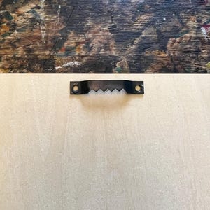 May include: A close-up of a light beige wooden surface with a black metal hanging hook attached to the top.
