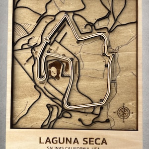 May include: A wooden wall art piece featuring a 3D relief map of the Laguna Seca racetrack in Salinas, California. The design includes raised track outlines, numbered turns, a compass rose, and the text Laguna Seca Salinas California, USA.