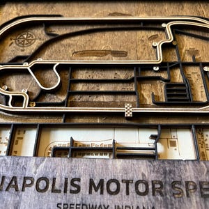 May include: A wooden 3D model of the Indianapolis Motor Speedway race track. The track is made of light brown wood and is surrounded by a dark brown wood frame. The track is detailed with black lines and white dots. The words "Indianapolis Motor Speedway" are engraved in the bottom of the frame.