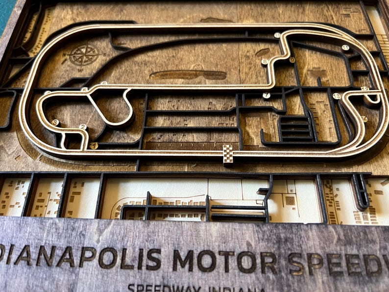 May include: A wooden 3D model of the Indianapolis Motor Speedway race track. The track is made of light brown wood and is surrounded by a dark brown wood frame. The track is detailed with black lines and white dots. The words "Indianapolis Motor Speedway" are engraved in the bottom of the frame.