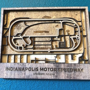 May include: A wooden wall art piece depicting the Indianapolis Motor Speedway race track. The track is outlined in black and the surrounding area is a light brown wood. The text "Indianapolis Motor Speedway" and "Speedway, Indiana" is printed below the track.