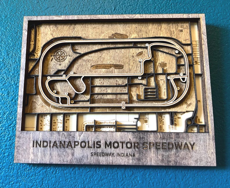 May include: A wooden wall art piece depicting the Indianapolis Motor Speedway race track. The track is outlined in black and the surrounding area is a light brown wood. The text "Indianapolis Motor Speedway" and "Speedway, Indiana" is printed below the track.