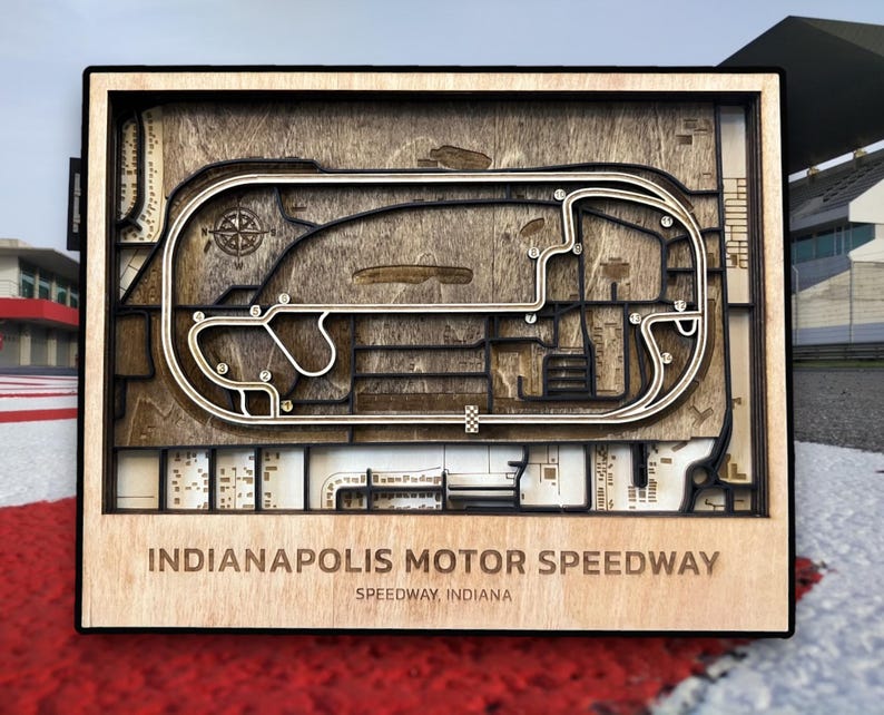 May include: A wooden wall art piece depicting the Indianapolis Motor Speedway race track. The track is outlined in black and the interior is a wood grain pattern. The text "Indianapolis Motor Speedway" and "Speedway, Indiana" is printed below the track.