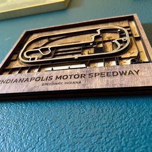May include: A wooden wall art piece featuring a 3D laser-cut design of the Indianapolis Motor Speedway race track. The text "Indianapolis Motor Speedway, Speedway, Indiana" is engraved below the track.
