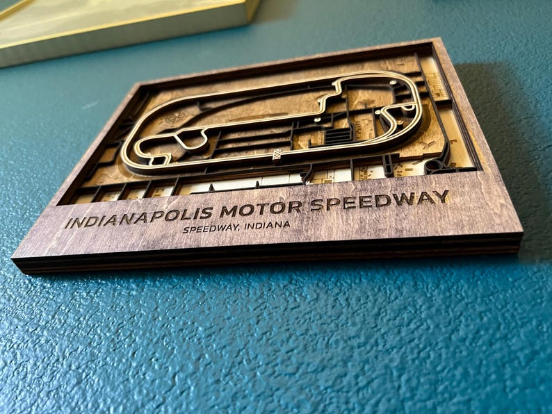 May include: A wooden wall art piece featuring a 3D laser-cut design of the Indianapolis Motor Speedway race track. The text "Indianapolis Motor Speedway, Speedway, Indiana" is engraved below the track.