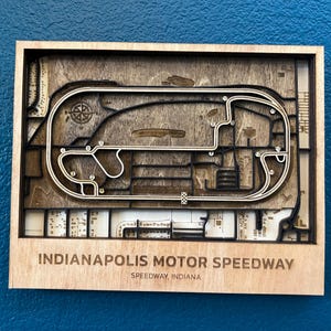 May include: A wooden wall art piece depicting the Indianapolis Motor Speedway race track. The track is outlined in black and the surrounding area is made of wood. The text "Indianapolis Motor Speedway" and "Speedway, Indiana" is printed below the track.