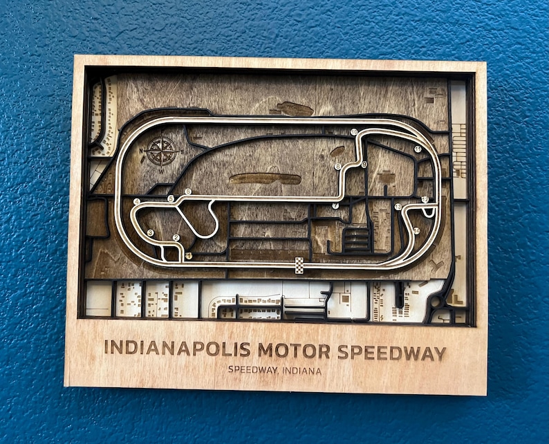 May include: A wooden wall art piece depicting the Indianapolis Motor Speedway race track. The track is outlined in black and the surrounding area is made of wood. The text "Indianapolis Motor Speedway" and "Speedway, Indiana" is printed below the track.