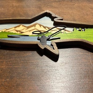 K&ouml;nnte beinhalten: Ein Wandbild aus Holz in Form eines Fisches mit einem geschichteten Design. Der Fisch ist mit einer Landschaftszene aus Bergen, B&auml;umen und einem Fluss bemalt. Eine Silhouette einer Person beim Angeln ist im Fluss zu sehen.