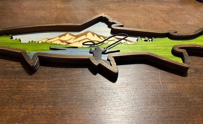 K&ouml;nnte beinhalten: Ein Wandbild aus Holz in Form eines Fisches mit einem geschichteten Design. Der Fisch ist mit einer Landschaftszene aus Bergen, B&auml;umen und einem Fluss bemalt. Eine Silhouette einer Person beim Angeln ist im Fluss zu sehen.