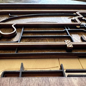 May include: A wooden race track model with a checkered flag finish line. The track is made of dark wood and has a light wood border. The track is raised off the surface and has a 3D effect.