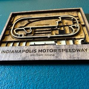 May include: A wooden wall art piece depicting the Indianapolis Motor Speedway race track. The track is made of layered wood and is surrounded by a dark brown frame. The text "Indianapolis Motor Speedway" and "Speedway, Indiana" is printed below the track.